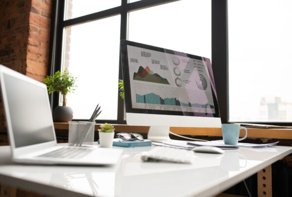 Workplace of analyst Computer monitor with graphical information on its screen, laptop, mug and other supplies on desk by office window